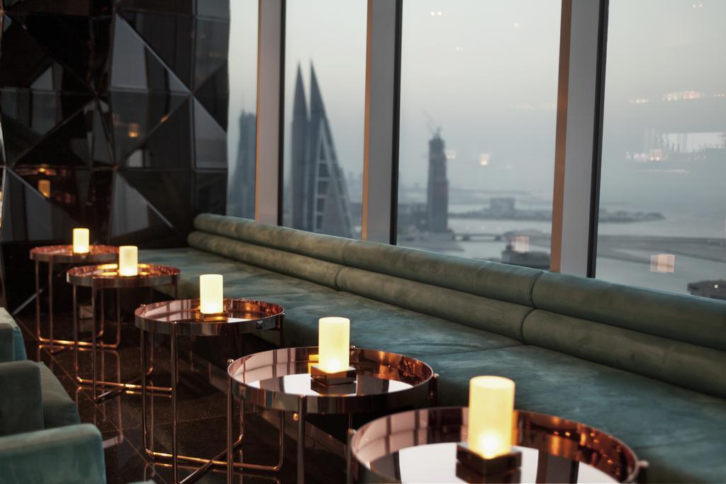The Domain Bahrain Hotel And Spa - Adults Friendly 16 Years Plus Манама Екстер'єр фото The photo depicts a stylish lounge area with a modern aesthetic. There are several circular tables made of metallic materials, each topped with a small glowing candle. A long, plush seating area in a teal color runs along the window, offering a view 
